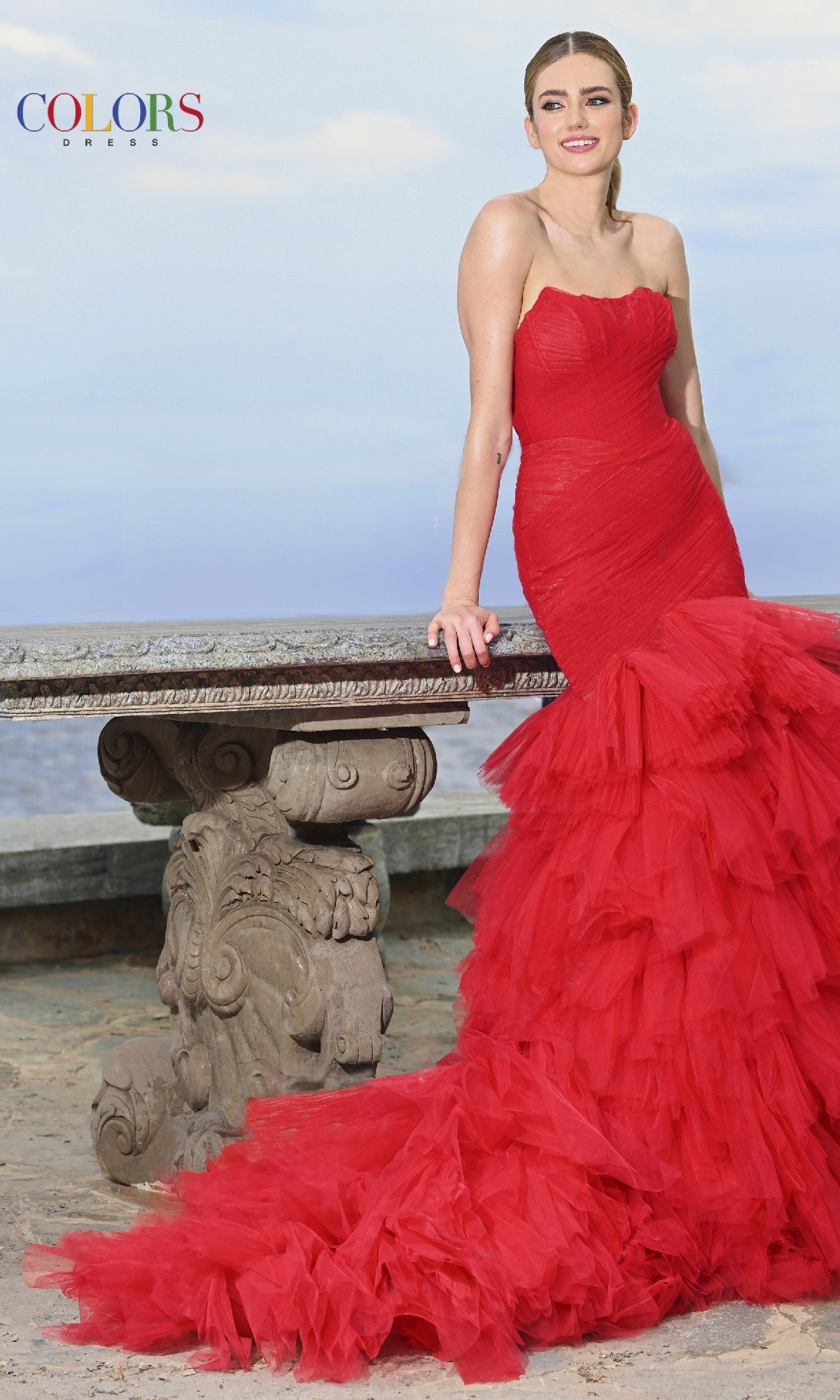 A woman wearing the Strapless Mermaid Prom Gown: Colors Dress 3631 with ruffled red layers leans against a stone bench outdoors, framed by the sky and ocean.